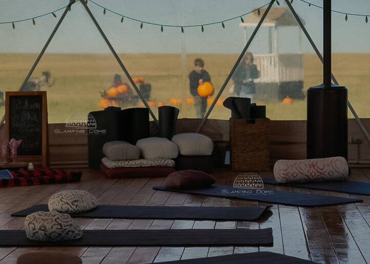 Interior of a 32-foot (10-meter) geodesic dome tent structure, sold by Glamping Dome Store, with a light khaki PVC cover, installed at Healing Grounds by Keys to Hope, Redcliff, Alberta, Canada. The dome is set up for a yoga or meditation session, featuring mats, cushions, and bolsters arranged on a wooden floor. The transparent panoramic windows provide views of an outdoor pumpkin patch and prairie landscape, creating a serene and inviting environment for relaxation and healing.