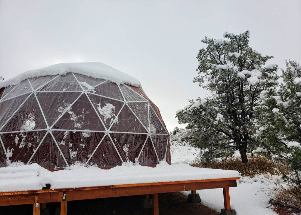 A geodesic dome tent in Arizona, USA, covered in fresh snow and set on a sturdy elevated wooden deck. Snow-laden trees surround the structure, showcasing its resilience in unexpected winter conditions. This image highlights Glamping Dome Store’s geodesic dome tents, designed to withstand wind and snow loads, ensuring a secure and comfortable all-season glamping experience—even in regions like Arizona.