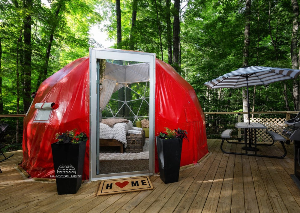 16.4-foot (5-meter) geodesic dome tent, sold by Glamping Dome Store, with a bold red PVC cover, set on a wooden deck in a tranquil forest setting at Little Red Homestead, a glamping destination in Lake Ann, Michigan, USA. The dome features a welcoming open glass door, a cozy interior visible through the entrance, and outdoor seating with a picnic table and umbrella, perfect for glamping retreats or eco-friendly outdoor living.