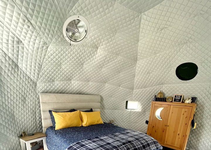 Interior of a geodesic dome tent Airbnb at Burnstick Lake Resort, Caroline, Alberta, Canada, showcasing a cozy bedroom with a stylish bed featuring yellow pillows and a navy comforter. The dome includes a solar exhaust fan for ventilation and a round clear PVC window with a flap closed from the outside. A mesh-screen vent is open, allowing fresh air circulation. Quilted insulation fabric lines the dome walls, ensuring comfort and warmth, making it ideal for unique outdoor accommodations or glamping setups.