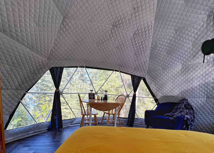 Interior of a geodesic dome tent Airbnb at Burnstick Lake Resort, Caroline, Alberta, Canada, a Glamping Dome Store project. Featuring quilted synthetic insulation fabric that ensures year-round comfort, it enhances warmth during colder months while keeping cool in summer. The space includes a blue armchair, a wooden dining table, and panoramic windows with curtains showcasing forest views. Perfect for eco-friendly glamping, Airbnb stays, and semi-permanent accommodations.