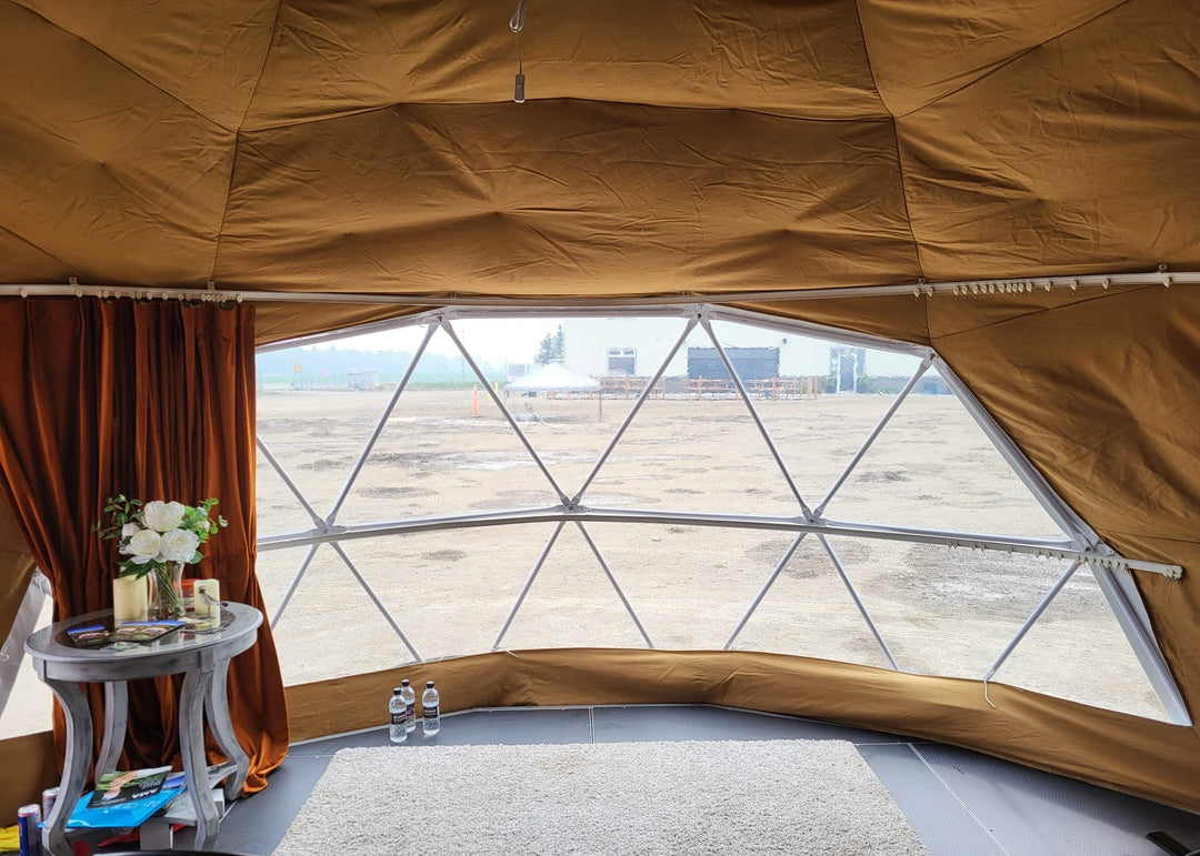 Interior of a geodesic dome tent by Glamping Dome Store, located in Rocky Mountain House, Alberta, Canada. Featuring bubble foil insulation fabric finished with brown canvas, a large clear PVC panoramic window with brown curtains, and cozy furnishings ideal for glamping setups or campground accommodations.