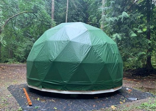 A green geodesic dome tent set up in a forested area, shown on a raised wooden platform for stability and protection.