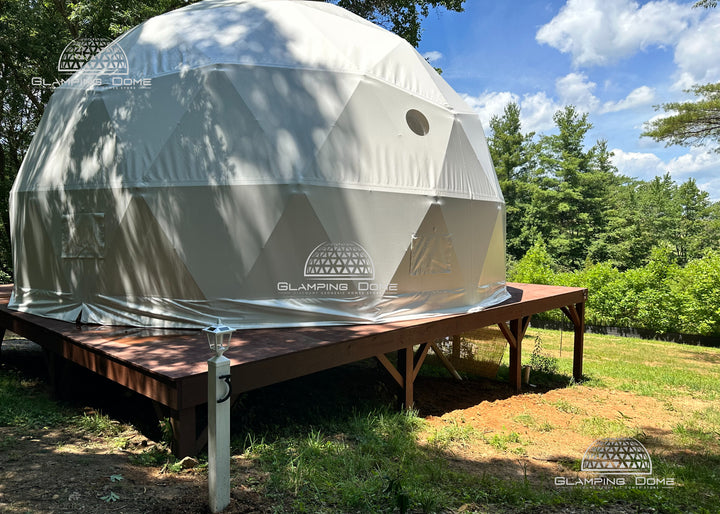 29.5-foot (9-meter) geodesic dome tent by Glamping Dome Store, installed at Big Little Clouds in Charlotte, North Carolina, USA. This dome features a durable white PVC cover and is elevated on a sturdy brown wooden platform, surrounded by lush greenery and blue skies. Designed for comfort and functionality, the dome includes two mesh-screen vents for adjustable airflow and a round clear PVC window, allowing ample daylight to illuminate the interior while maintaining proper ventilation.