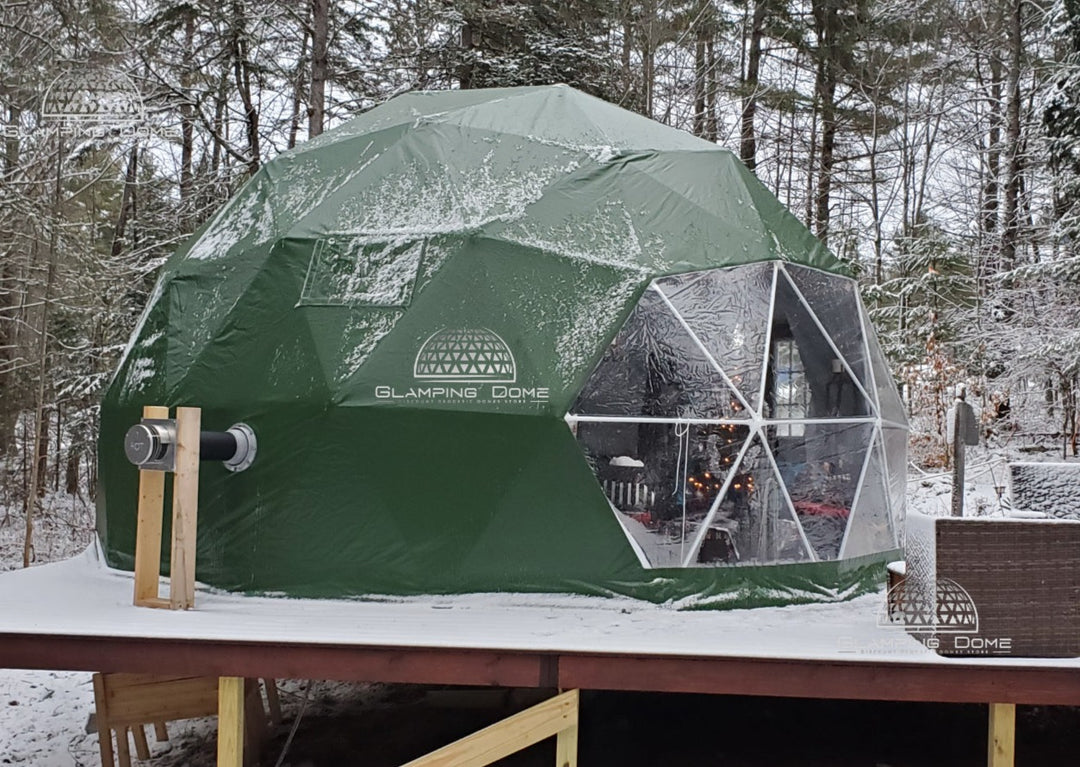 Geodesic dome tent, sold by Glamping Dome Store, located in Maine, New Hampshire, USA. The dome tent features a forest green PVC cover with a transparent panoramic window, set on a wooden platform in a snow-dusted winter forest. A wood stove pipe extends from the side, highlighting its all-season functionality. Inside, warm lighting creates a cozy and inviting atmosphere, ideal for glamping in colder climates.