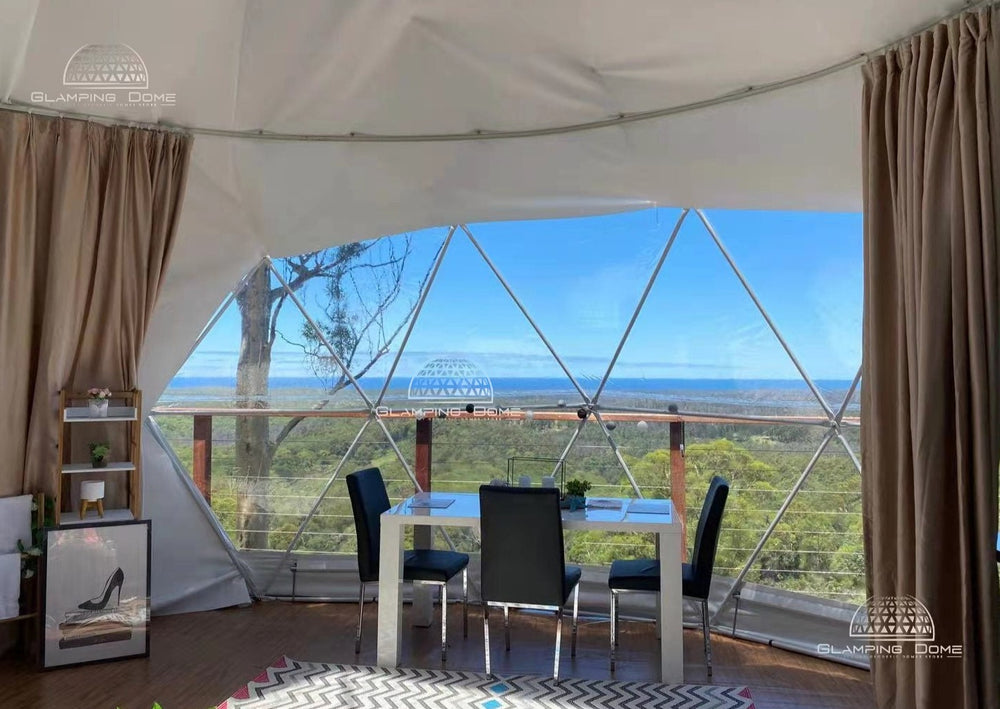 Interior view of a 23 ft (8 m) geodesic dome tent by Glamping Dome Store, showcasing a dining area with a panoramic window offering breathtaking views of lush greenery and distant horizons. The stylish setup includes a sleek white table, black chairs, and modern decor, creating an inviting atmosphere for dining or relaxation. The dome’s spacious interior and durable design make it ideal for glamping retreats, creative studios, or eco-friendly living.