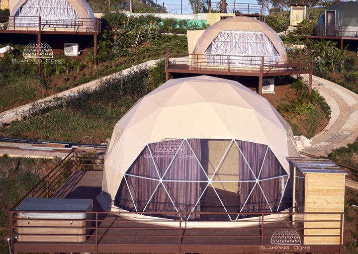 A serene glamping setup featuring multiple 23 ft (8 m) geodesic dome tents by Glamping Dome Store. Each dome is situated on individual elevated platforms, complete with private wooden decks and hot tubs, offering a luxurious retreat surrounded by lush greenery. The domes showcase panoramic windows with curtains for privacy, blending comfort and natural beauty. Ideal for ecotourism, luxury retreats, or unique vacation stays.