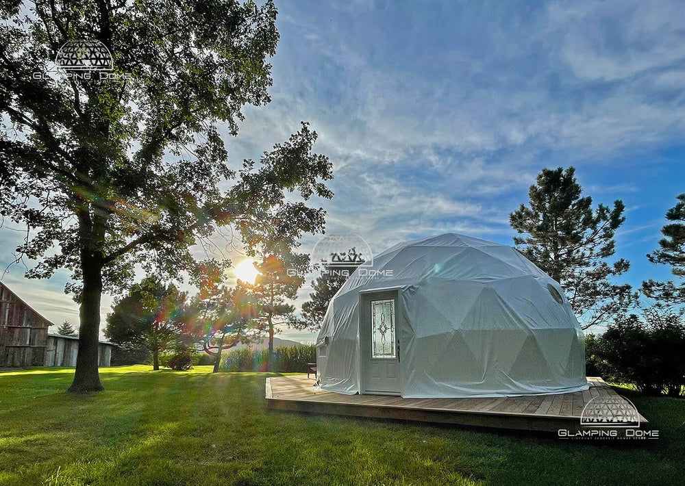 23-foot (7-meter) geodesic dome tent, a project by Glamping Dome Store, located at Maison des Prés with Dome & Jacuzzi in Saint-Basile-le-Grand, Quebec, Canada. The dome features a weather-resistant white PVC cover, a stylish glass-panelled door, and is set on a wooden platform surrounded by lush greenery. This tranquil retreat offers an idyllic glamping experience.