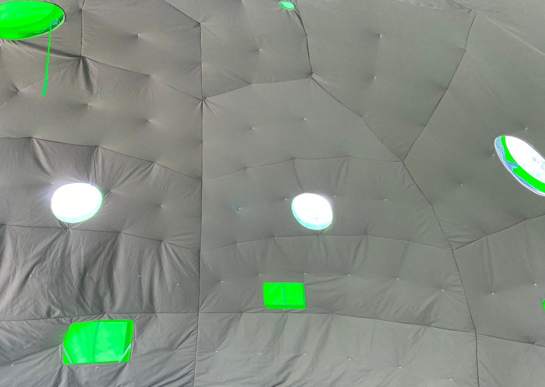 Interior of a geodesic dome tent by Glamping Dome Store, featuring off-white quilted synthetic insulation fabric with reflective bubble foil and canvas finish. The dome includes three round clear PVC windows for natural light, two mesh-screen vents, and an opening for a solar exhaust fan. The PVC cover is visible in lime green from the outside. Project located in Sylvan Lake, Alberta, Canada, ideal for geodesic dome tent buyers and glamping enthusiasts.